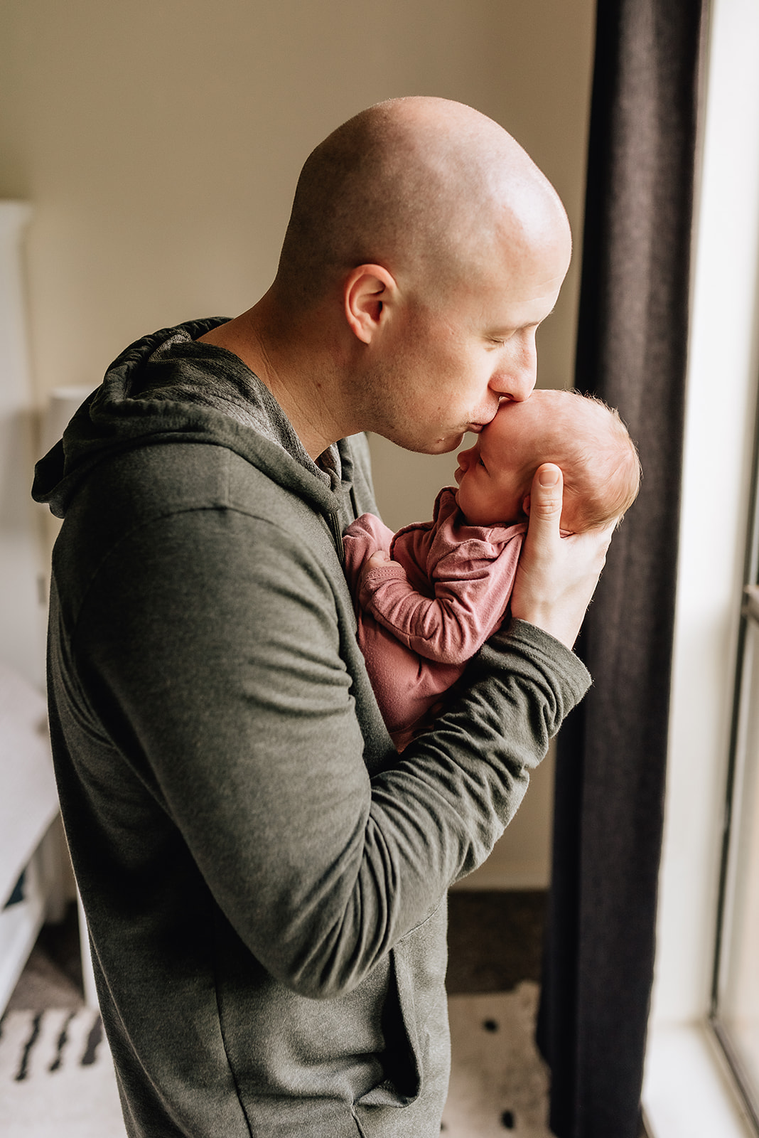 A father stands in front of a window and kisses the forehead of his newborn daughter