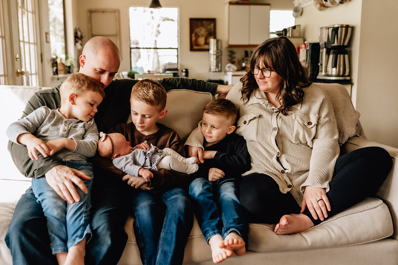 A family of six sit on a tan couch in their home together with everyone focused on their newborn baby in the middle North Houston Birth Center