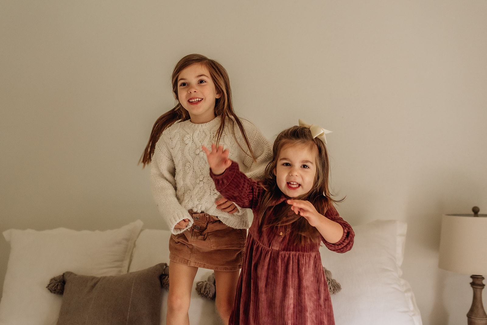 Two sisters jump and play on a bed in a red dreess and a knit sweater Threadfare