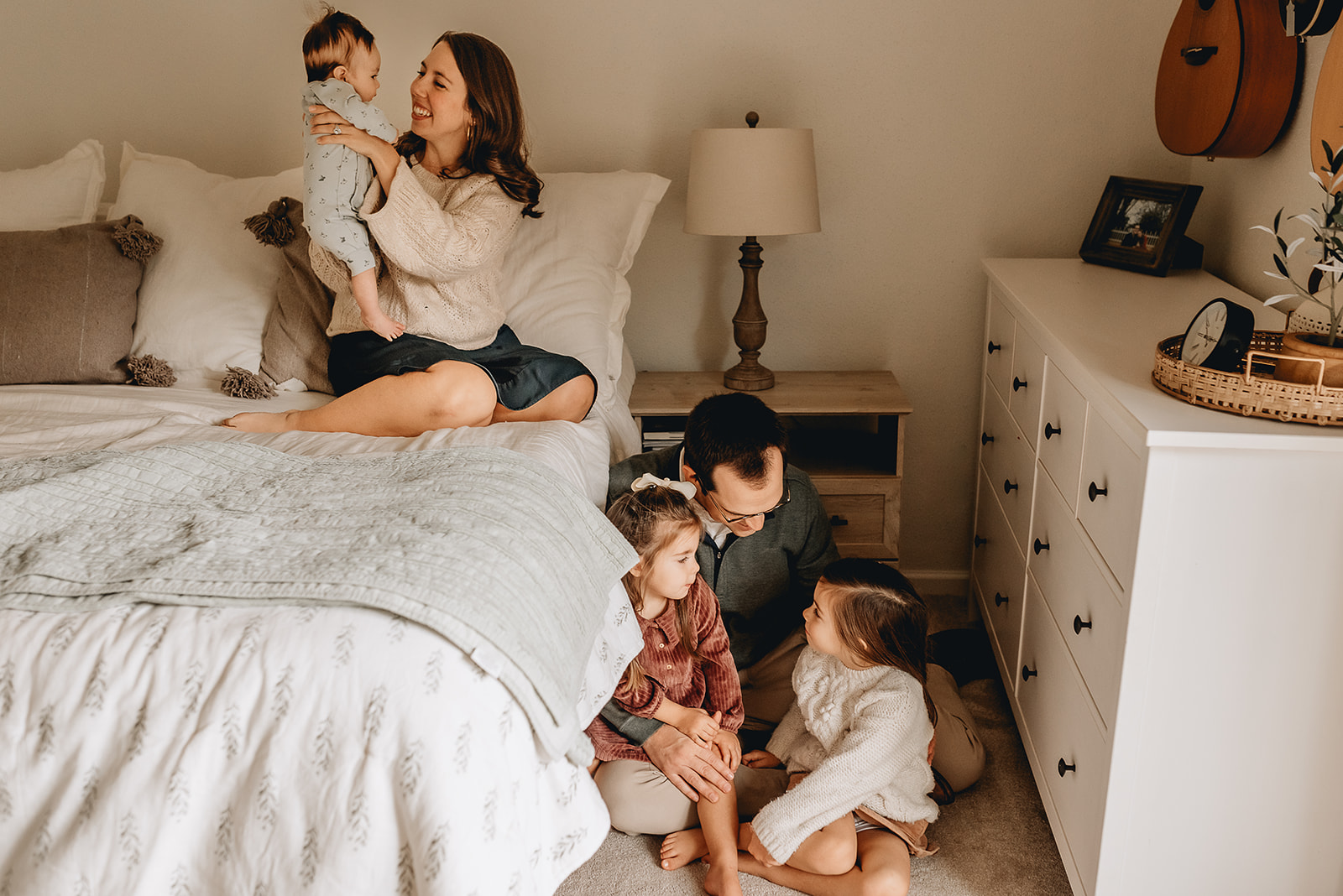 Mom sits on a bed playing with her newborn son in the air while dad sits on the floor next to the bed playing with their two other daughters Threadfare