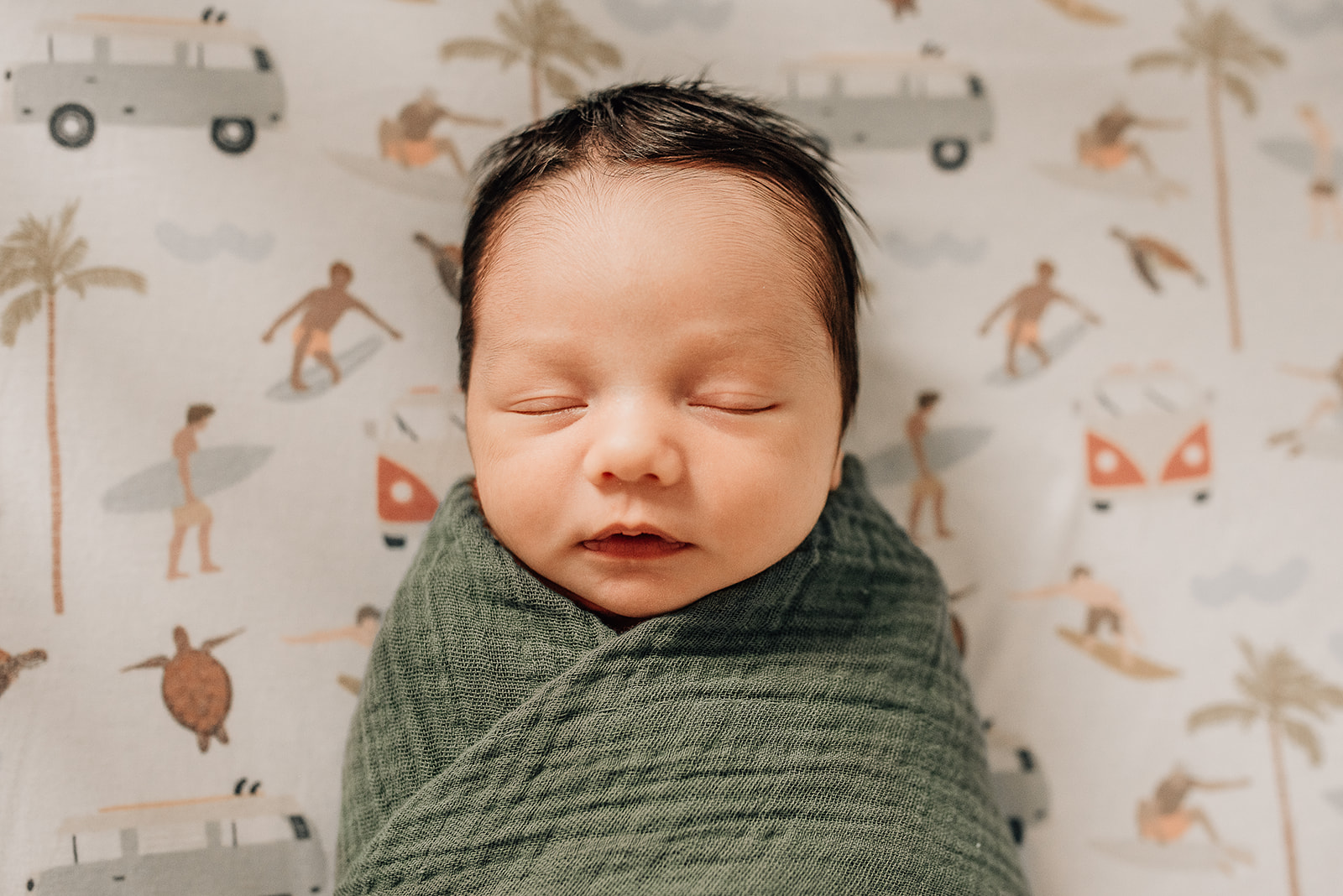 A newborn baby sleeps in a green swaddle on surfer sheets covered crib