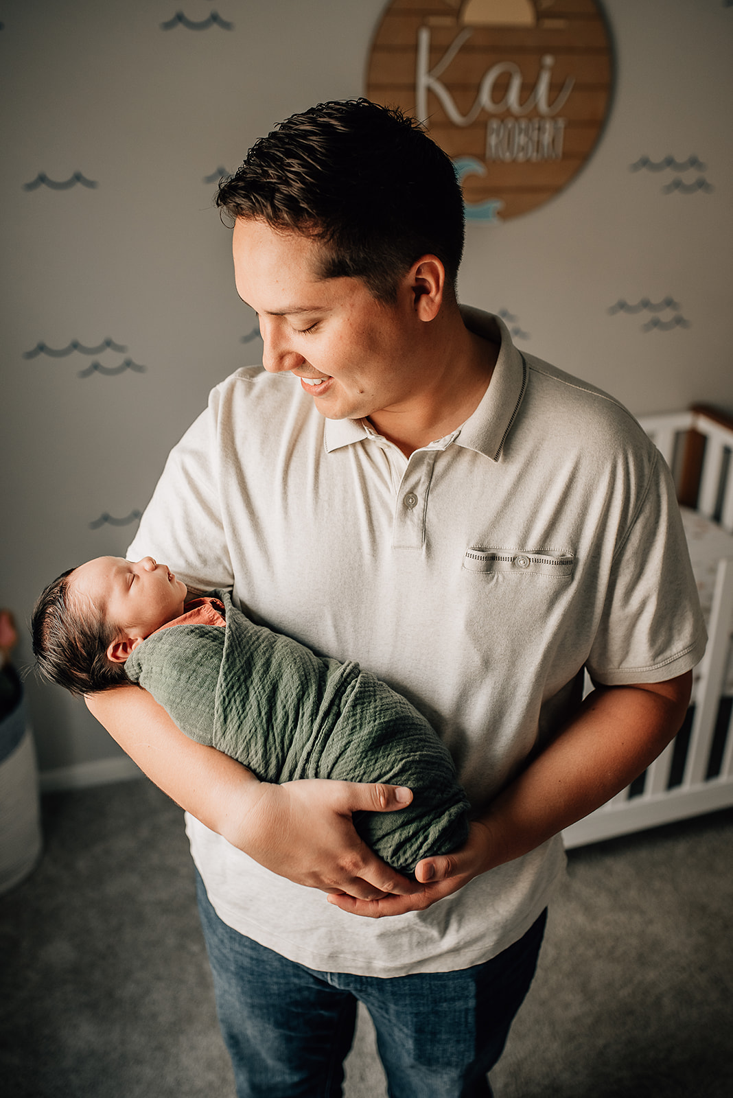 Dad wears a grey polo while holding his newborn son sleeping in a green swaddle in a nursery Sanna Baby