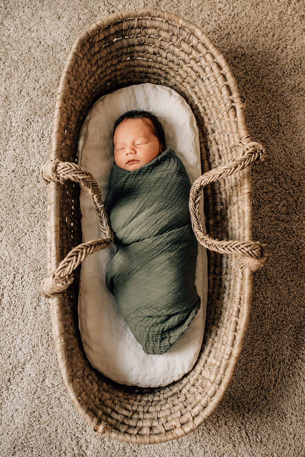 A newborn baby sleeps in a long woven basket in a green swaddle on a carpet floor Sanna Baby