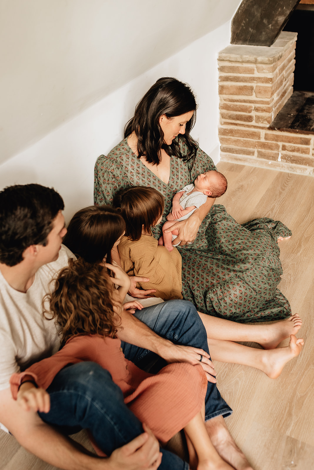 A mother in a green dress lits on the floor next to a fireplace while Dad and their three daughters sit next to her looking in