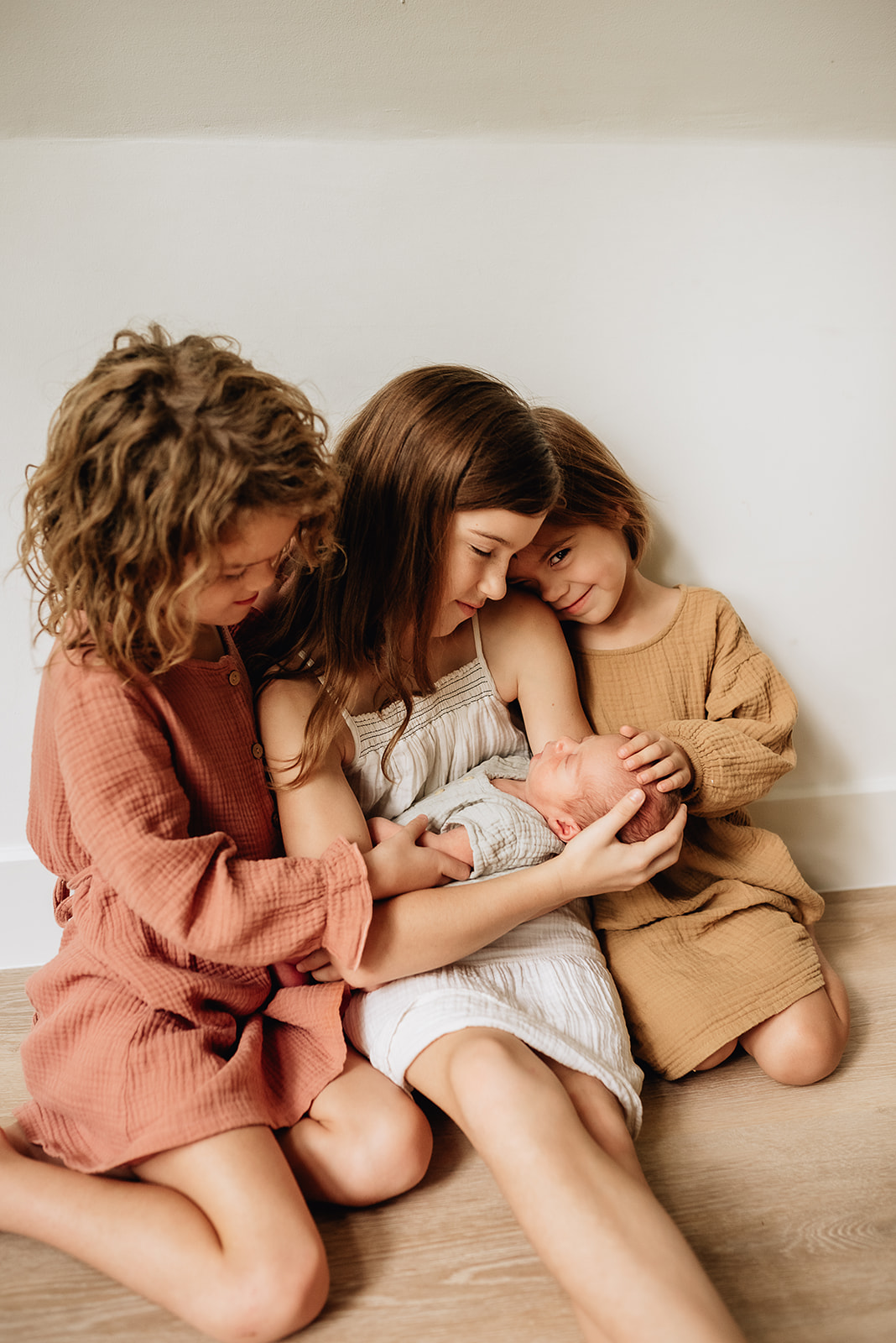 Three sisters sit on the floor against a wall holding their newborn baby sibling Baby and Kids 1st