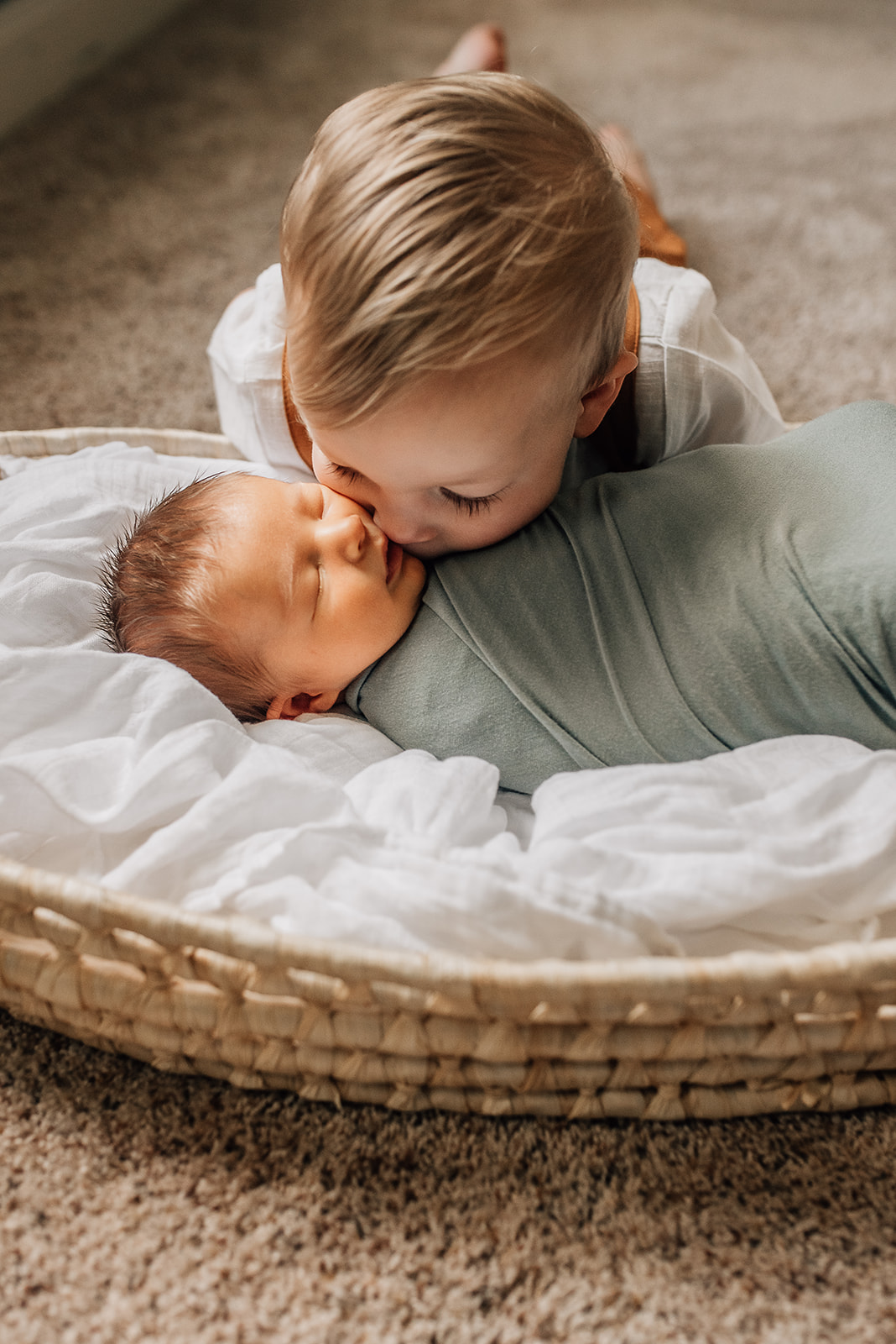 A young boy kisses the cheek of his newborn baby sibling while it sleeps in a basket Cy Fair Birth Center