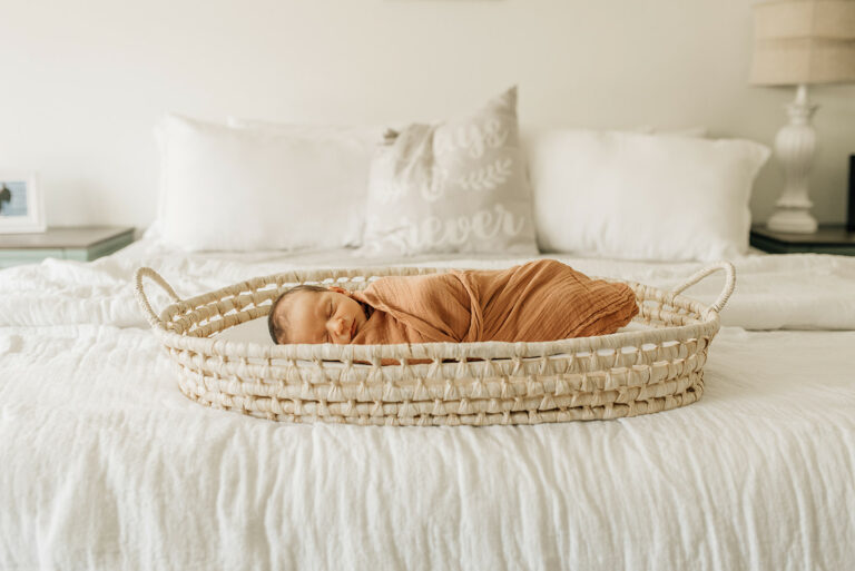 A newborn baby sleeps in a woven basket on a white bed while swaddled Houston baby shower venues