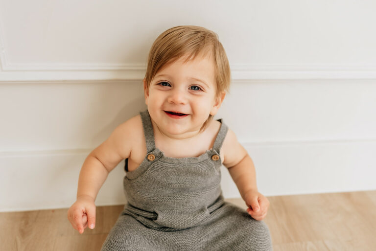 A toddler boy in knit grey overalls smiles and sits in a studio