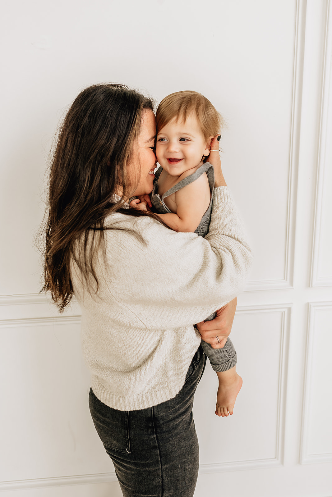 A mother snuggles her toddler son while standing in a studio in a white sweater