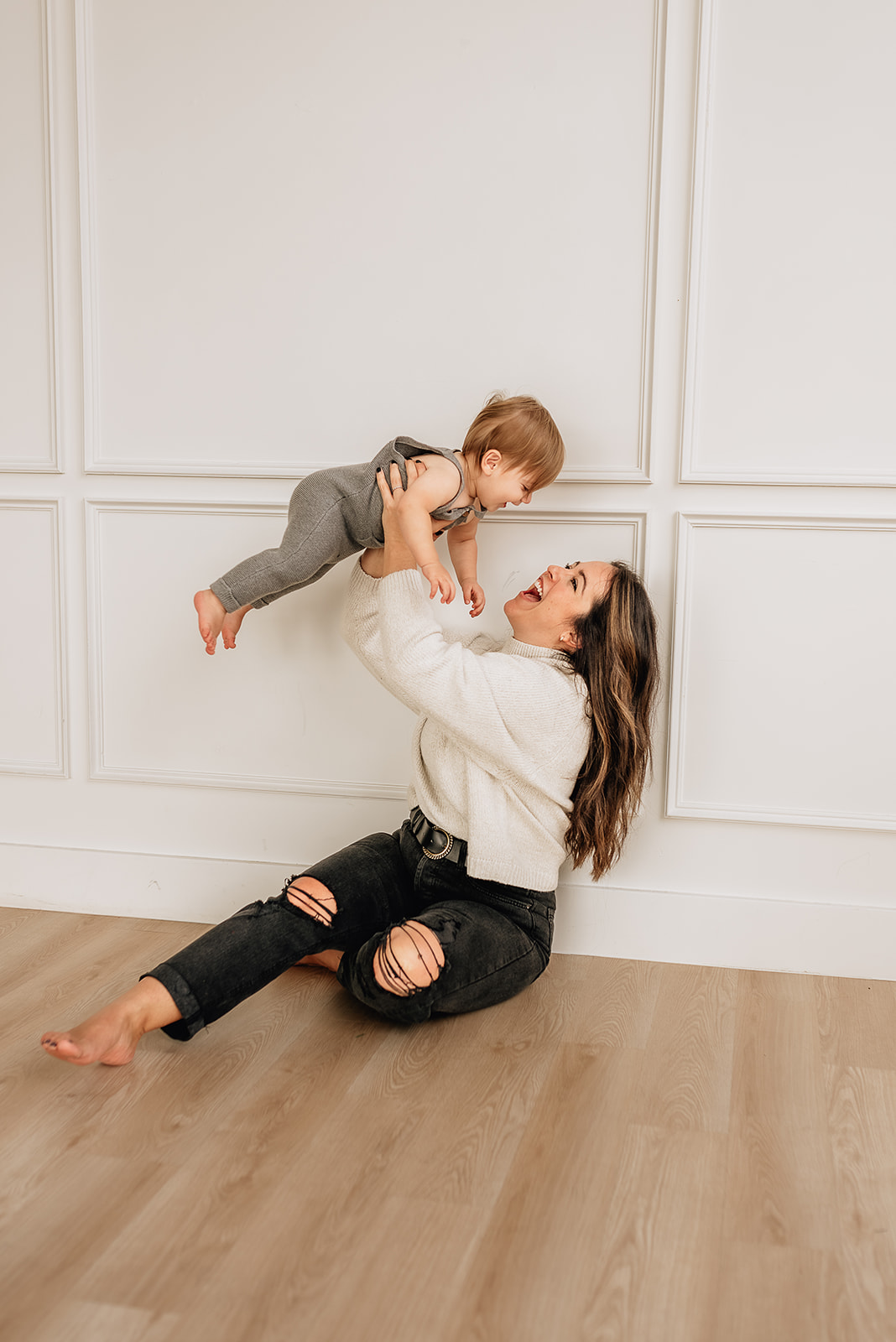 A mother in black pants and white sweater plays with her toddler son over her head while sitting on the floor of a studio houston midwives
