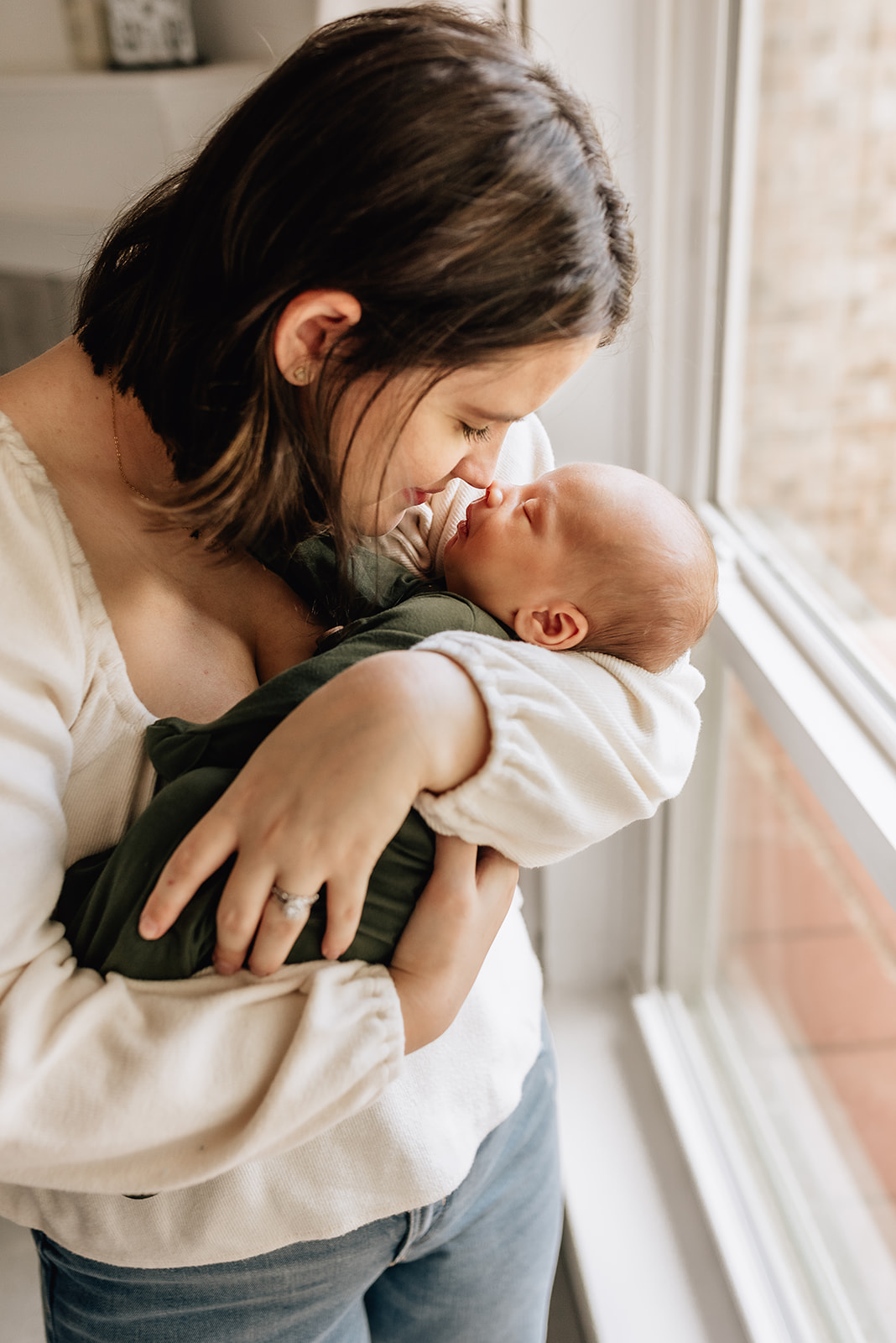 A mother in a white blouse boops her nose to her sleepiing newborn's nose while standing in a window midwife in the heights