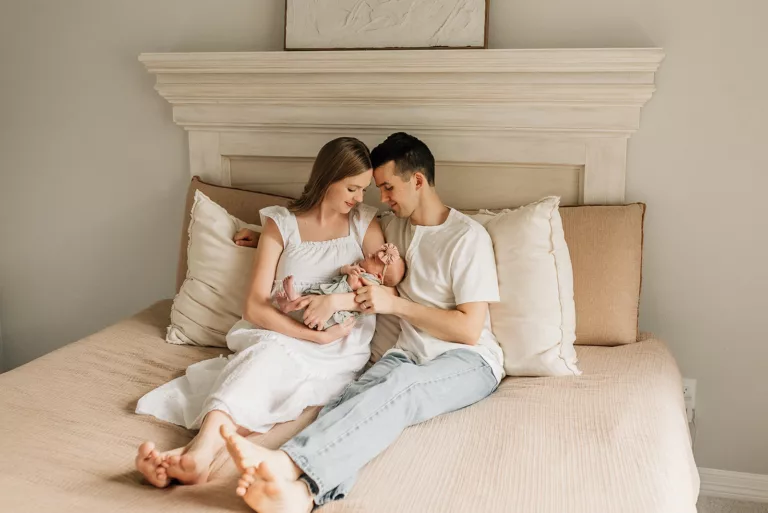 A mom and dad sit on a bed while their newborn baby lays in mom's arms