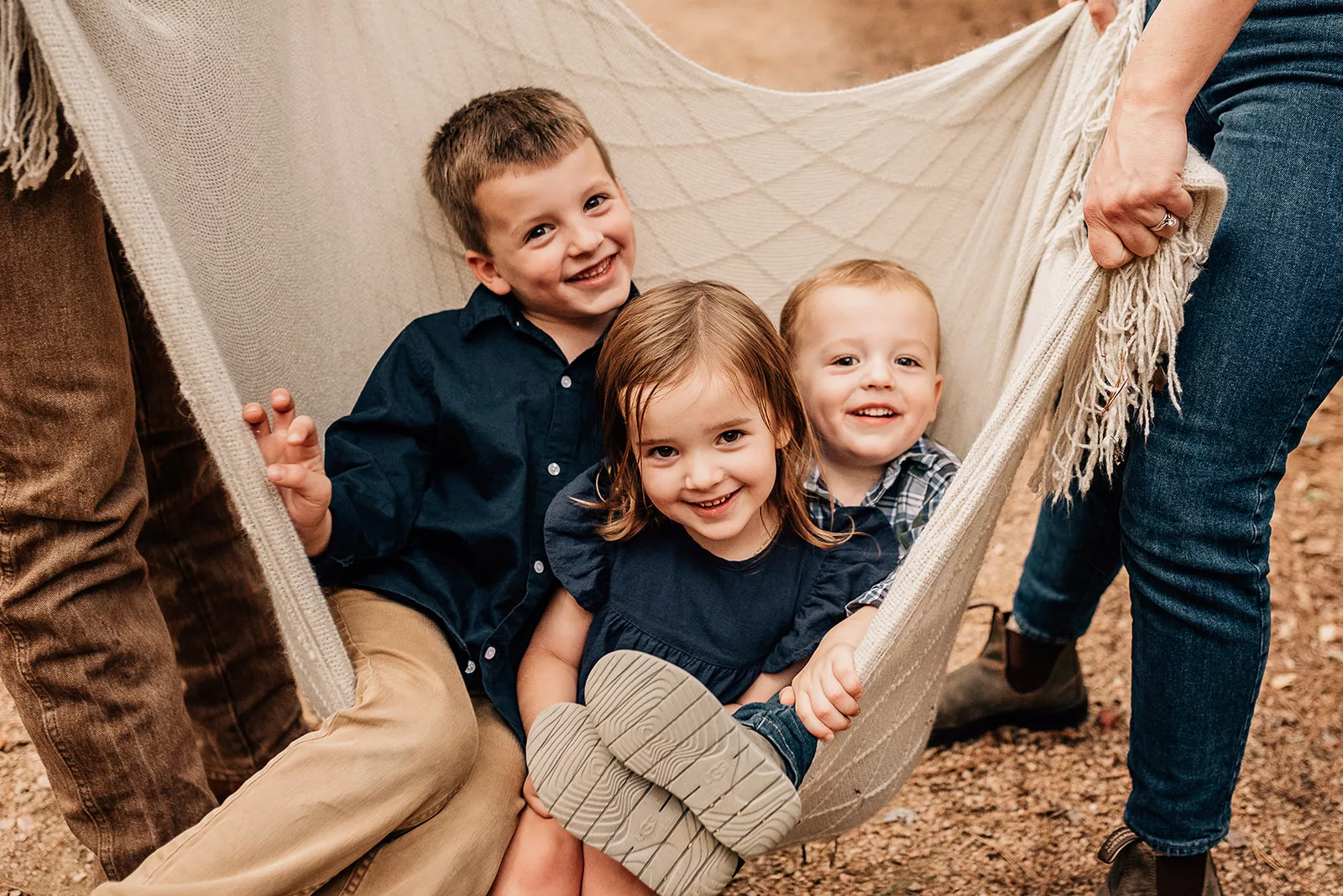 Three siblings sit in a picnic blanket like a hammock being held by mom and dad Houston Baby Boutiques