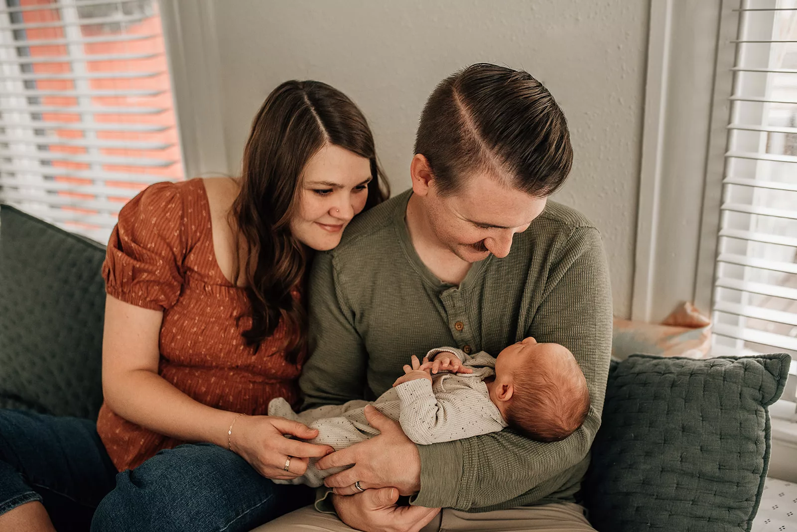 New parents sit together on a green couch looking down at their newborn baby in dad's arms