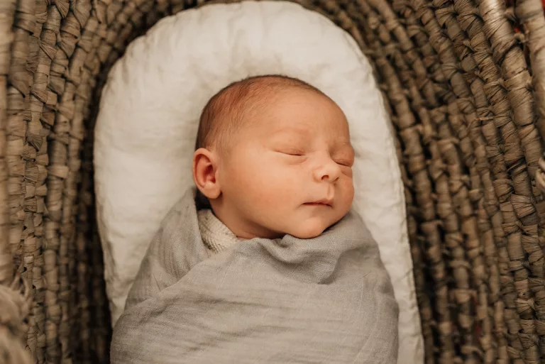 A newborn baby sleeps in a wicker basket in a grey blanket and swaddle Houston Lactation Consultant