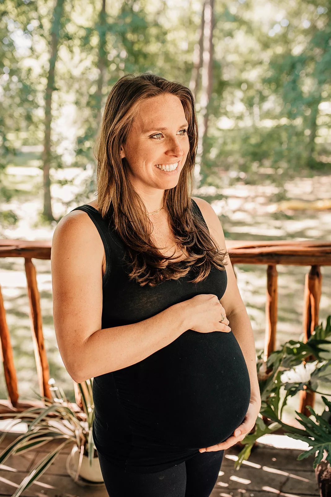 A mom to be stands on a wooden porch smiling in the sun and holding her bump in a black maternity shirt