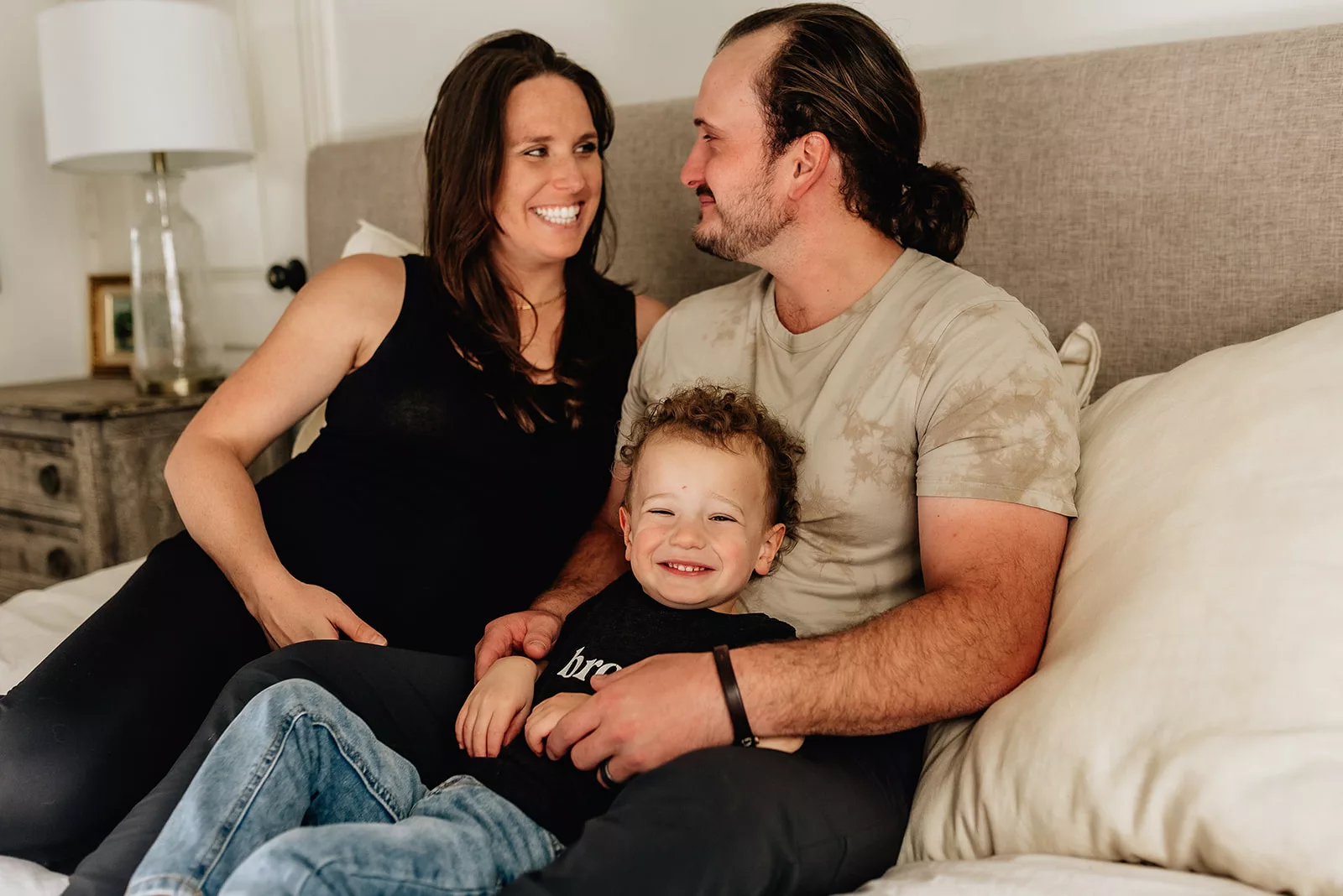 A pregnant mom smiles at her husband as they sit on a bed with their toddler son