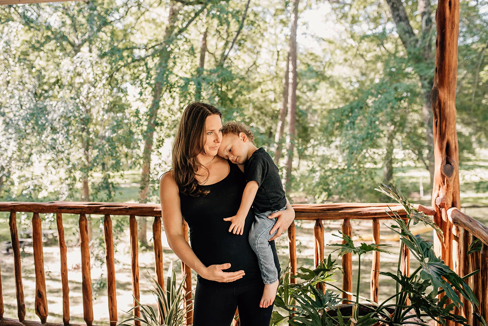 A pregnant mother stands on a porch holding her son on her hip as they both hold the bump