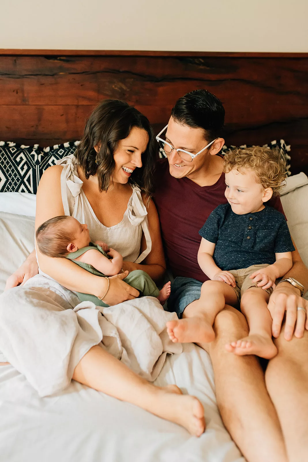 A family of four sit together on a bed together smiling with the newborn baby