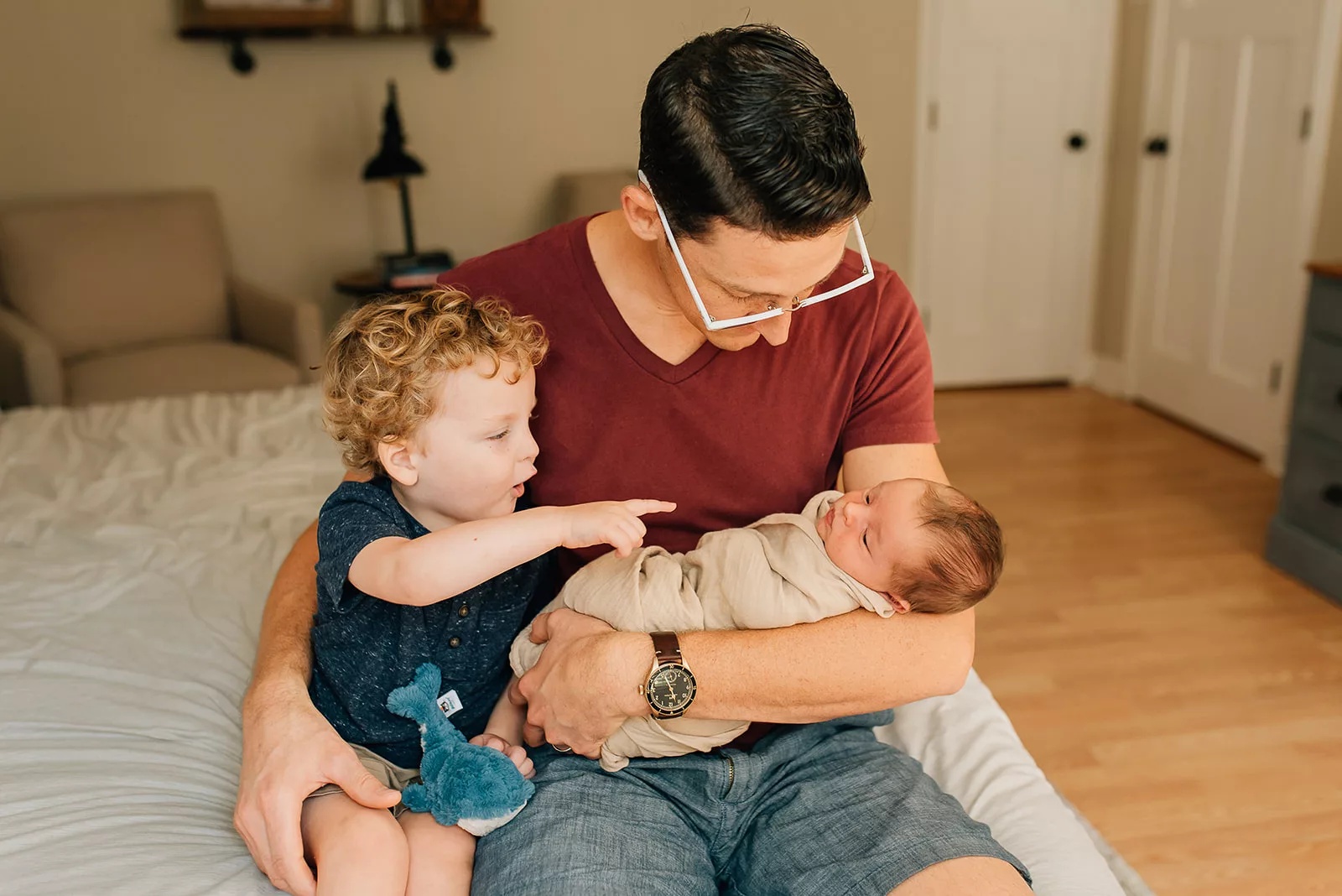 A father sits on the edge of the bed holding his newborn baby in his arms while playing with their toddler son before visiting Houston Pediatricians