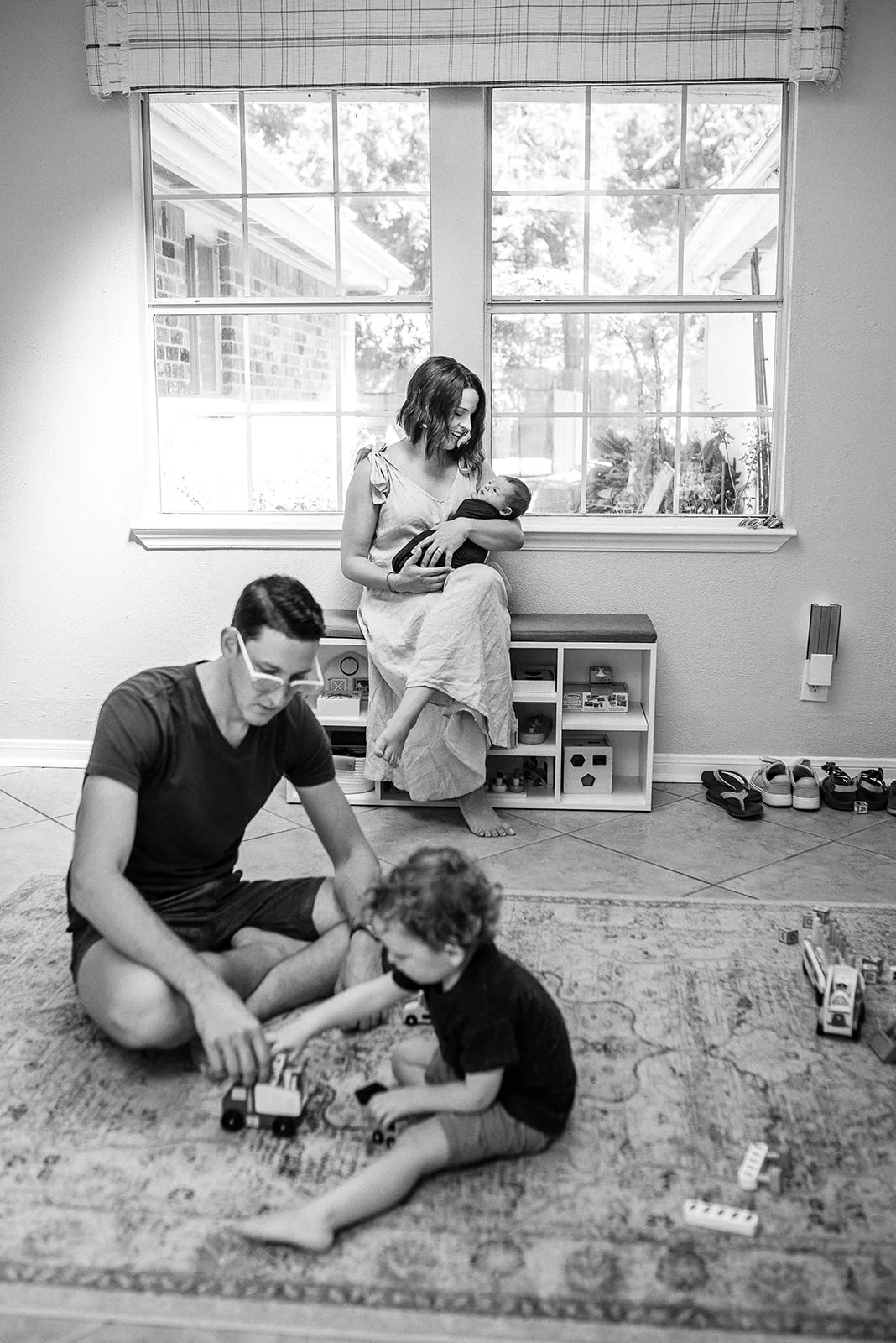 A mom sits on a toy bench holding her sleeping newborn baby while her husband plays with toys in the middle of the room with their toddler son after visiting Houston Pediatricians