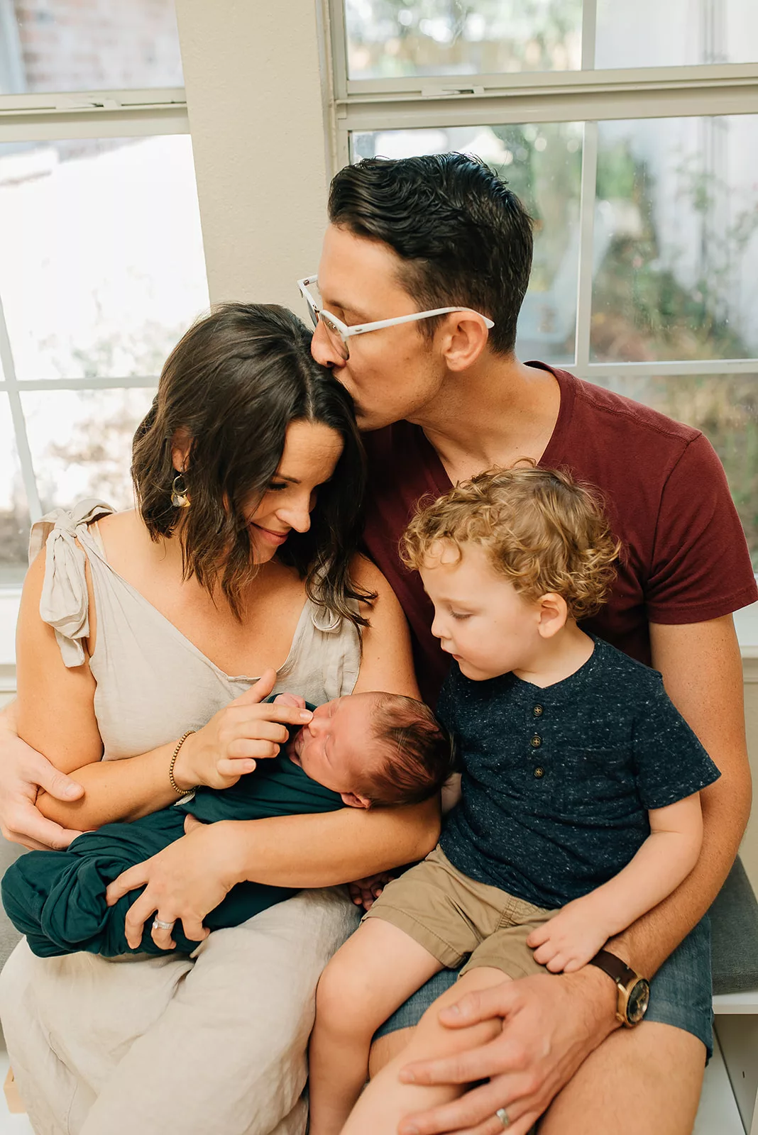 A mom touches the nose of her newborn baby in her arms while being kissed by her husband as their toddler son sits on his lap after visiting Houston Pediatricians