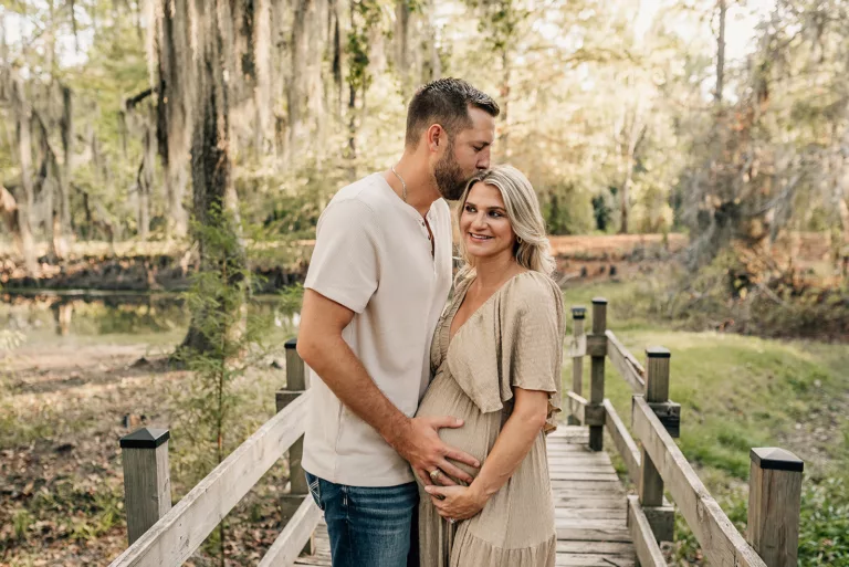 Expecting parents stand on a boardwalk as he kisses her head and holds the bump