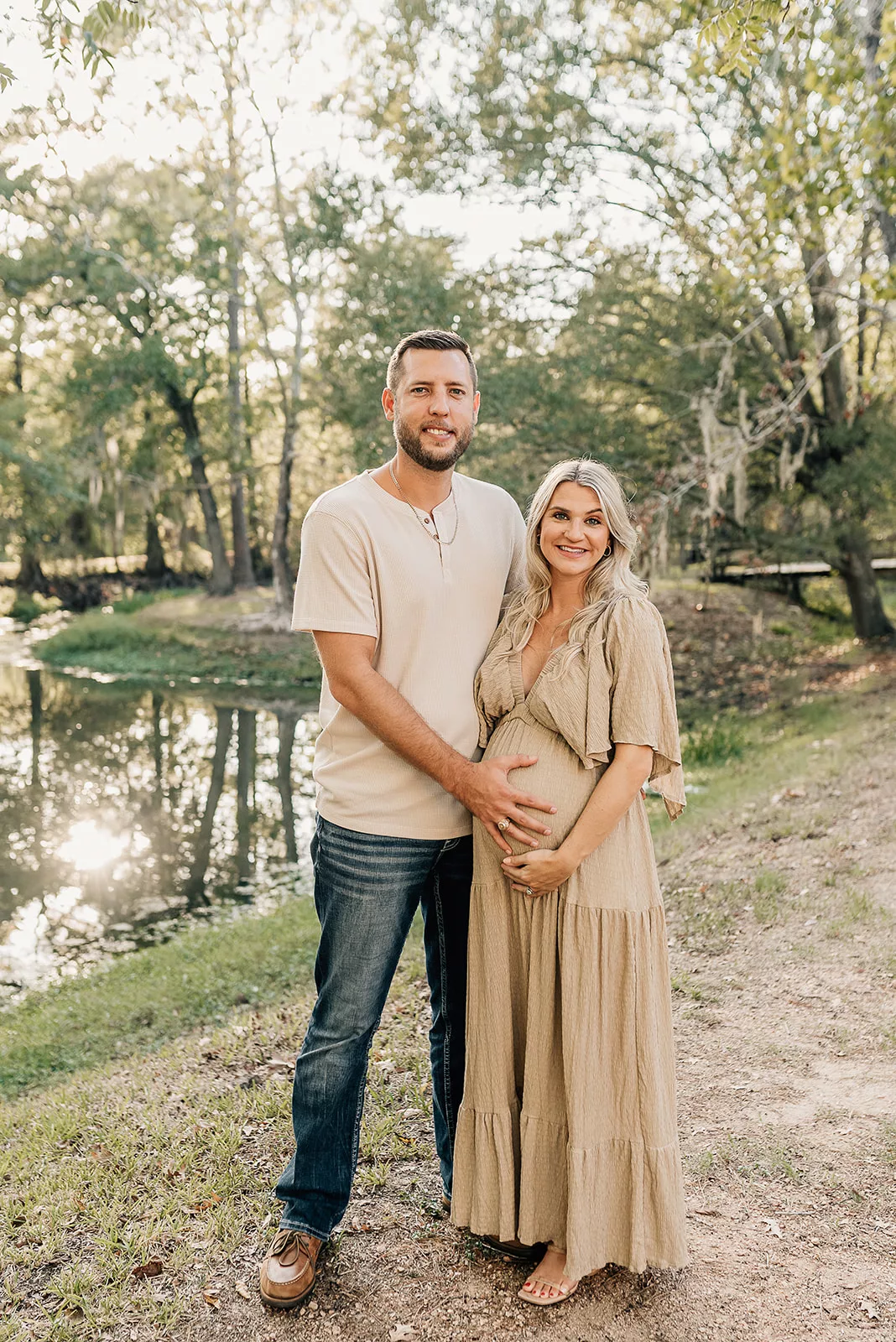 Expecting parents stand on the edge of a lake holding the bump