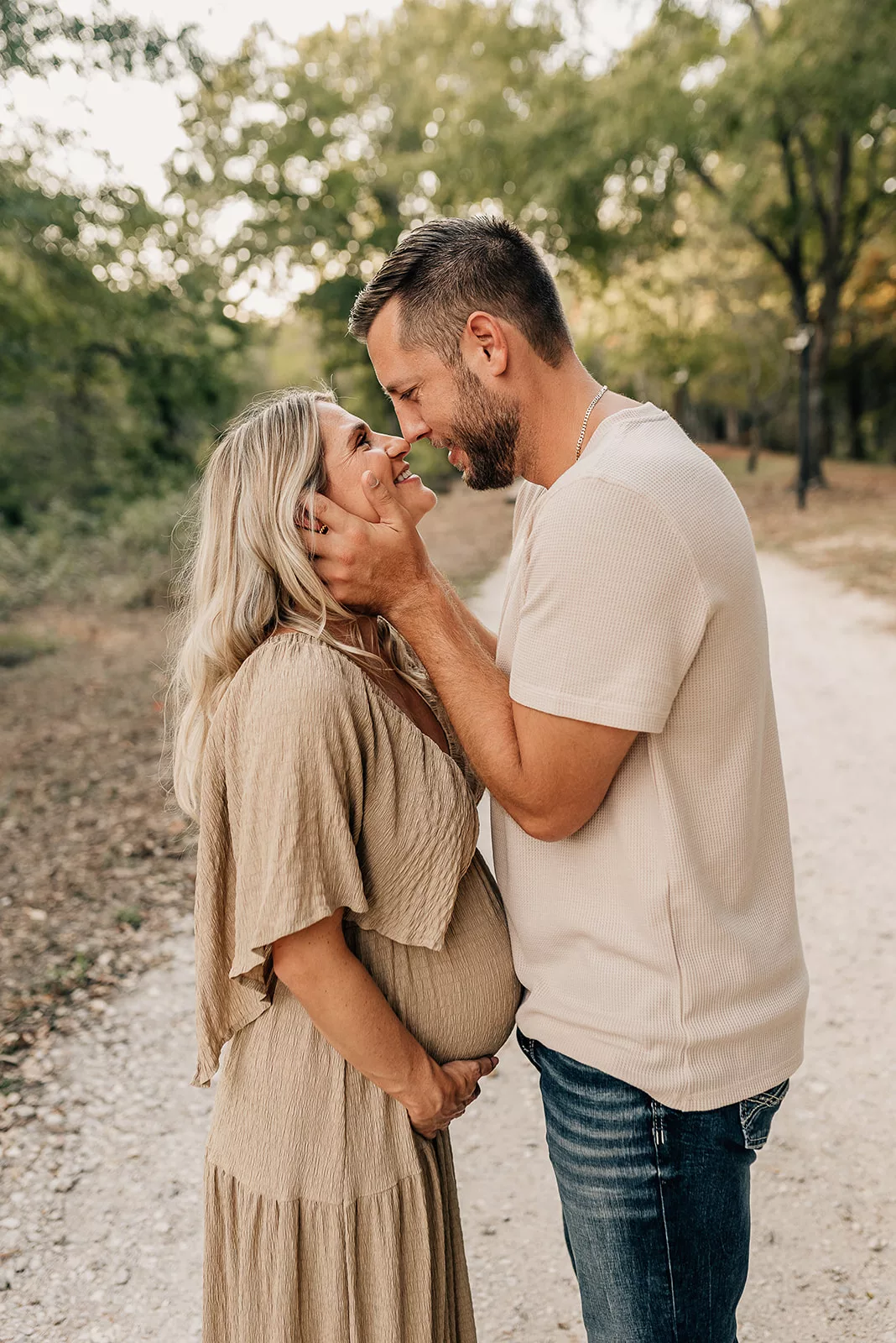 A mom to be touches noses with her husband as they stand in a gravel park path