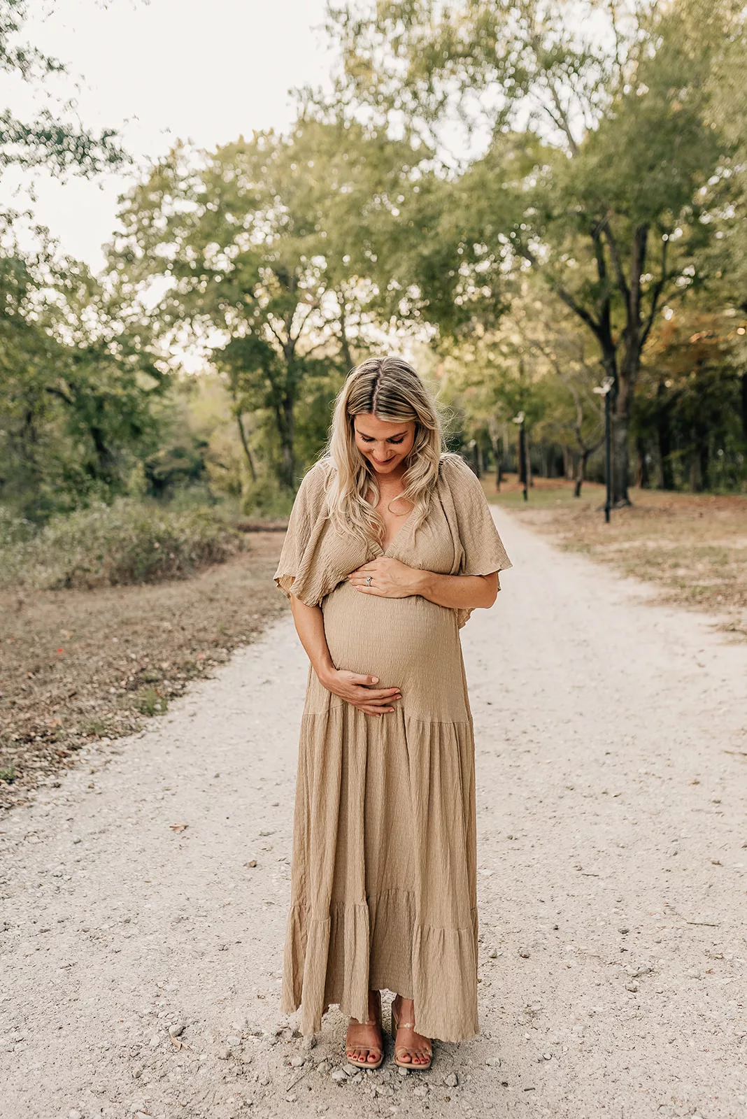 A mom to be stands in a park path smiling down to her bump in a tan dress after meeting with a Houston Postpartum Doula