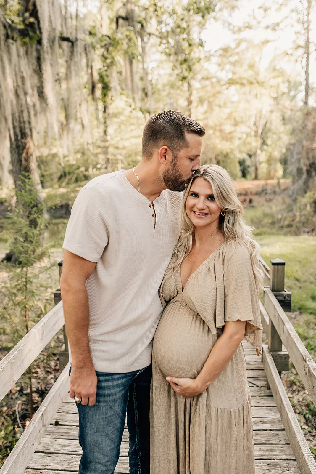 A mom to be stands on a wooden boardwalk holding her bump while her husband kisses her head