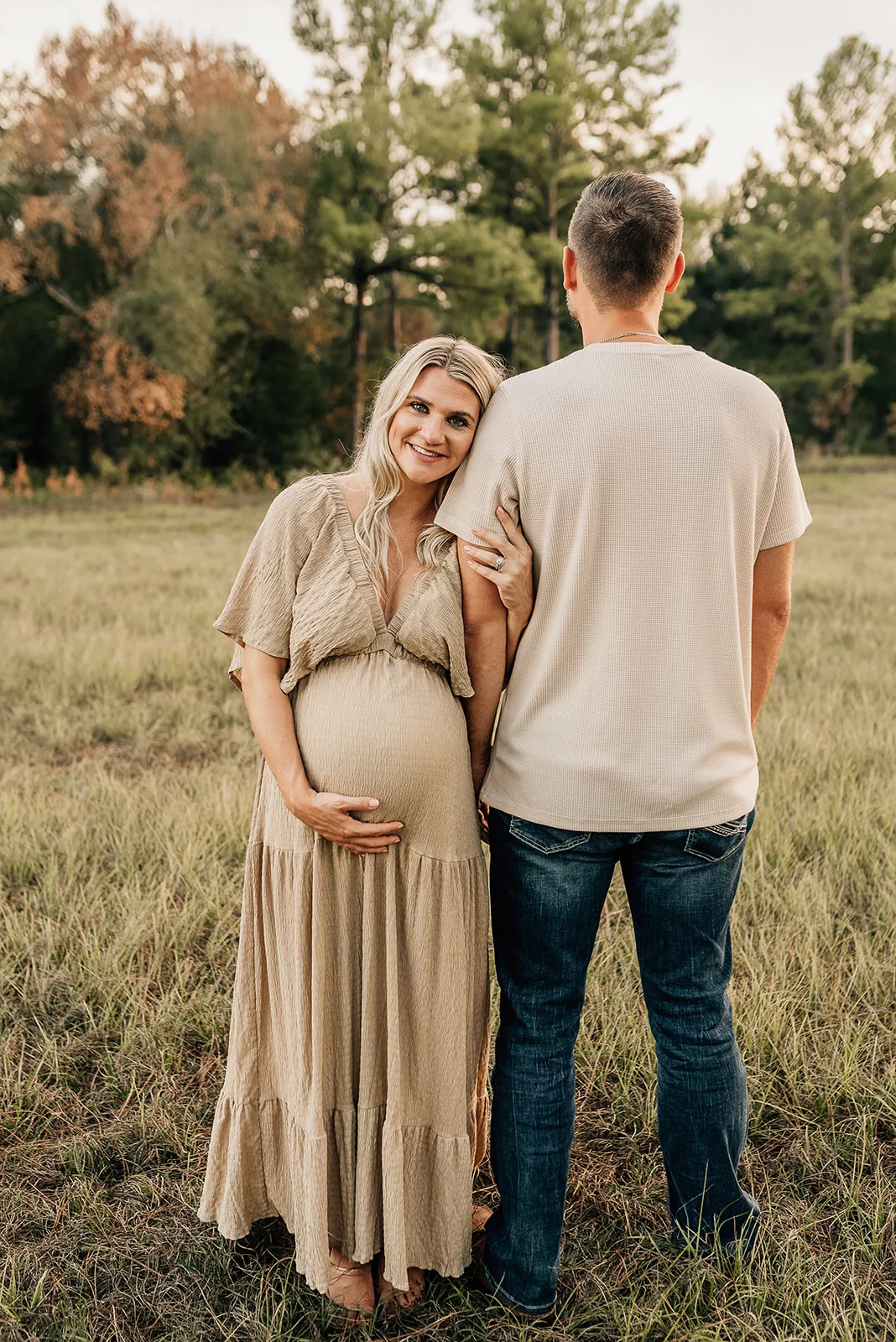 A mom to be leans on the shoulder of her husband as they stand in a field at sunset before visiting a Houston Postpartum Doula
