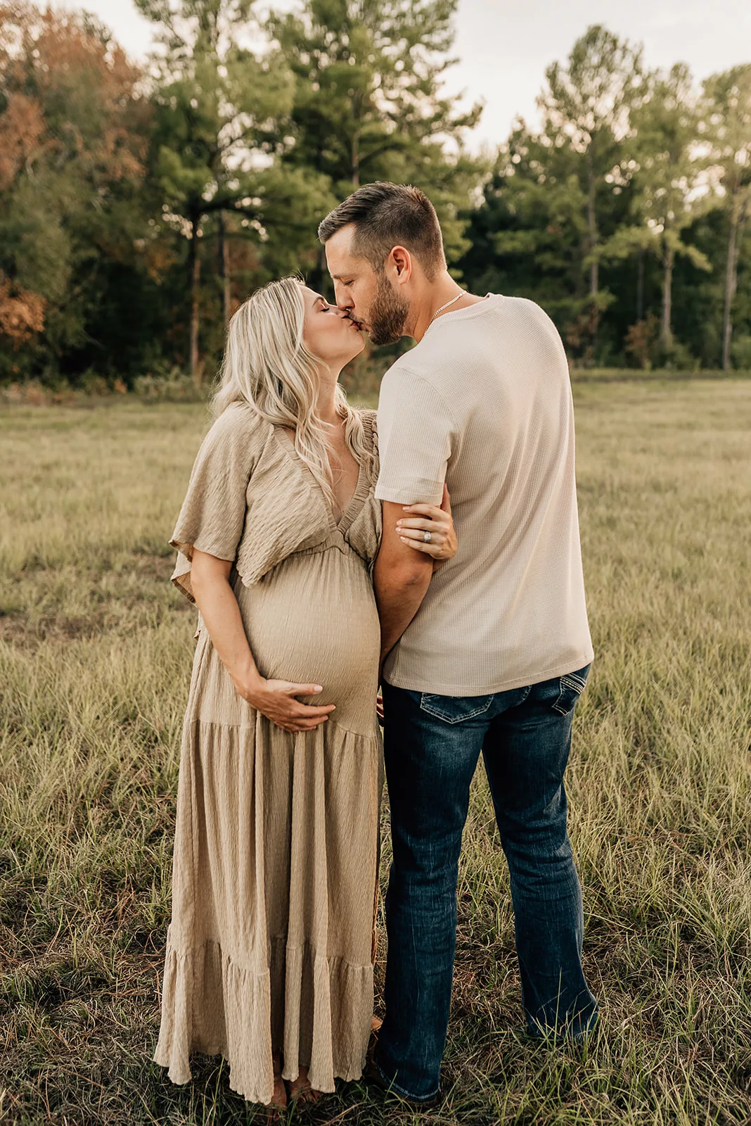 A mom to be holds onto the arm of her husband while they kiss in a field at sunset after meeting with a Houston Postpartum Doula