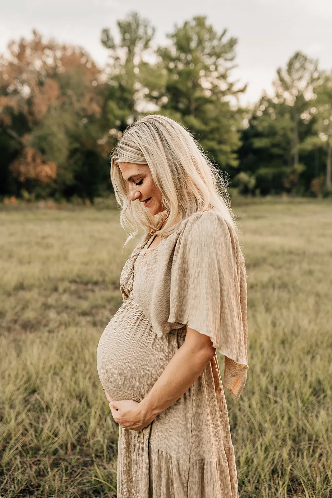A mom to be in a tan dress stands in a field smiling down at the bump in her hands before meeting with a Houston Postpartum Doula