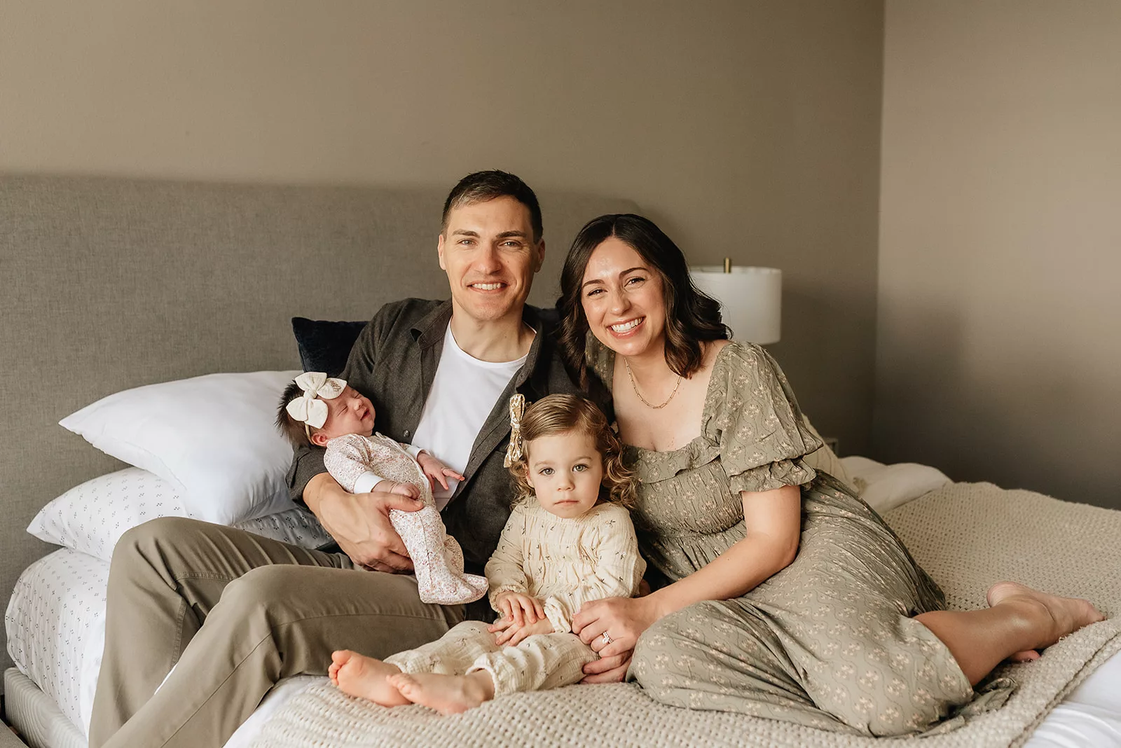 A mom and dad sit on a bed with their newborn baby and young toddler daughter