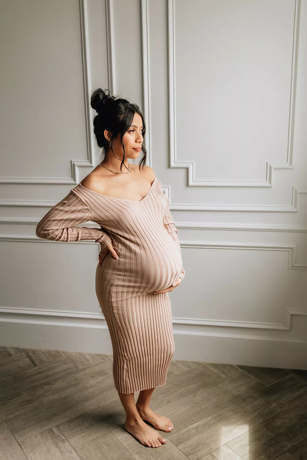 A mom to be in a beige maternity dress stands in a studio looking out the window and holding her bump after her 3D Ultrasound In Houston