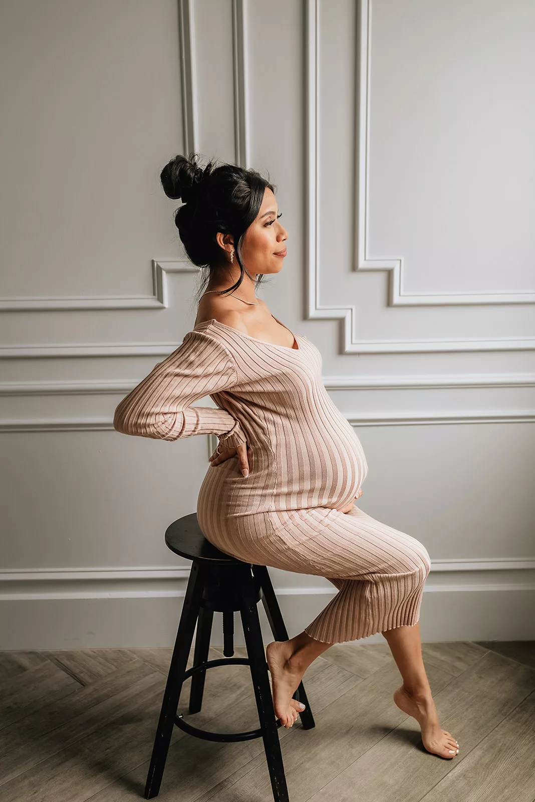 A mother to be in a beige maternity dress sits on a stool in a studio smiling out a window after getting her 3D Ultrasound In Houston