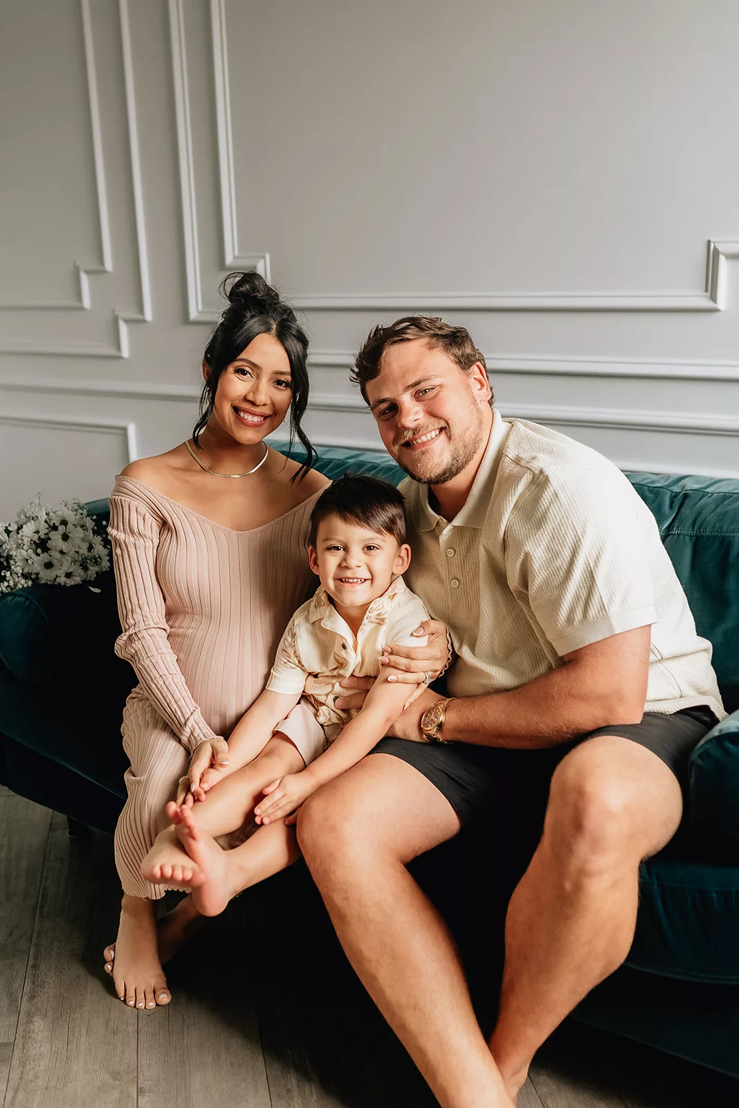 A pregnant mom and dad sit on a green couch smiling with their toddler son