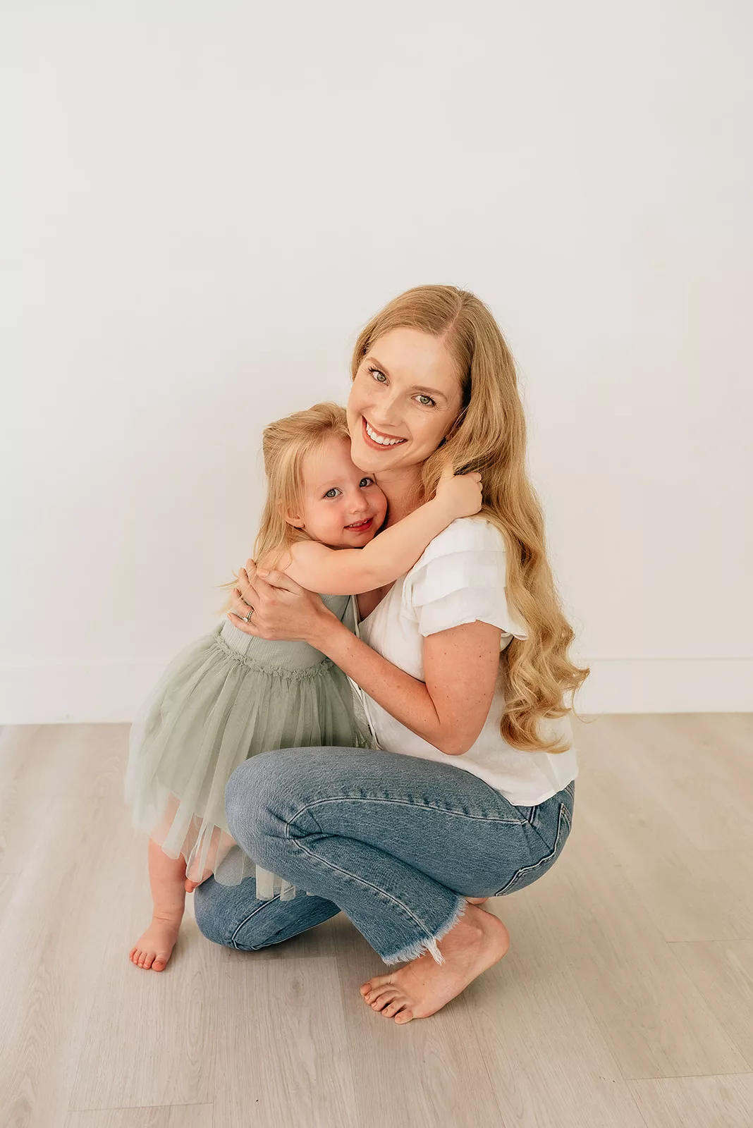 A mom in jeans kneels down to hug her toddler daughter in a green dress in a studio