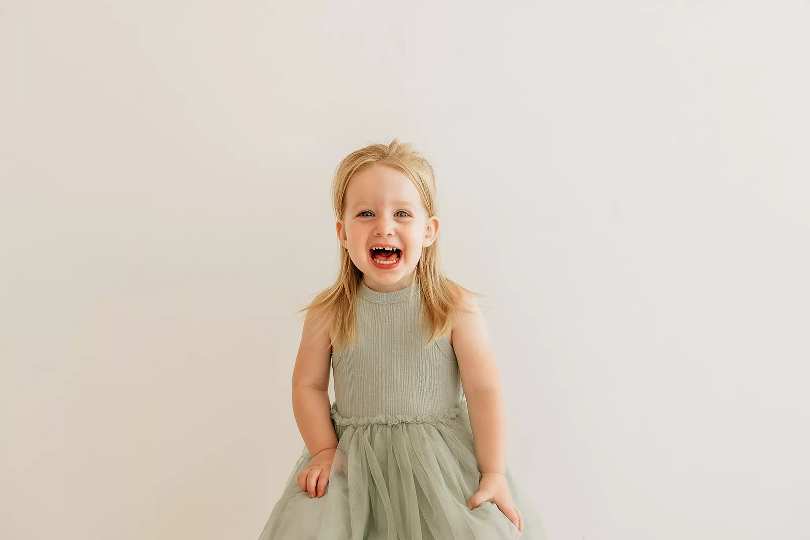 A toddler girl smiles big with mouth open while sitting on a stool in a studio in a green dress after visiting Houston Daycares