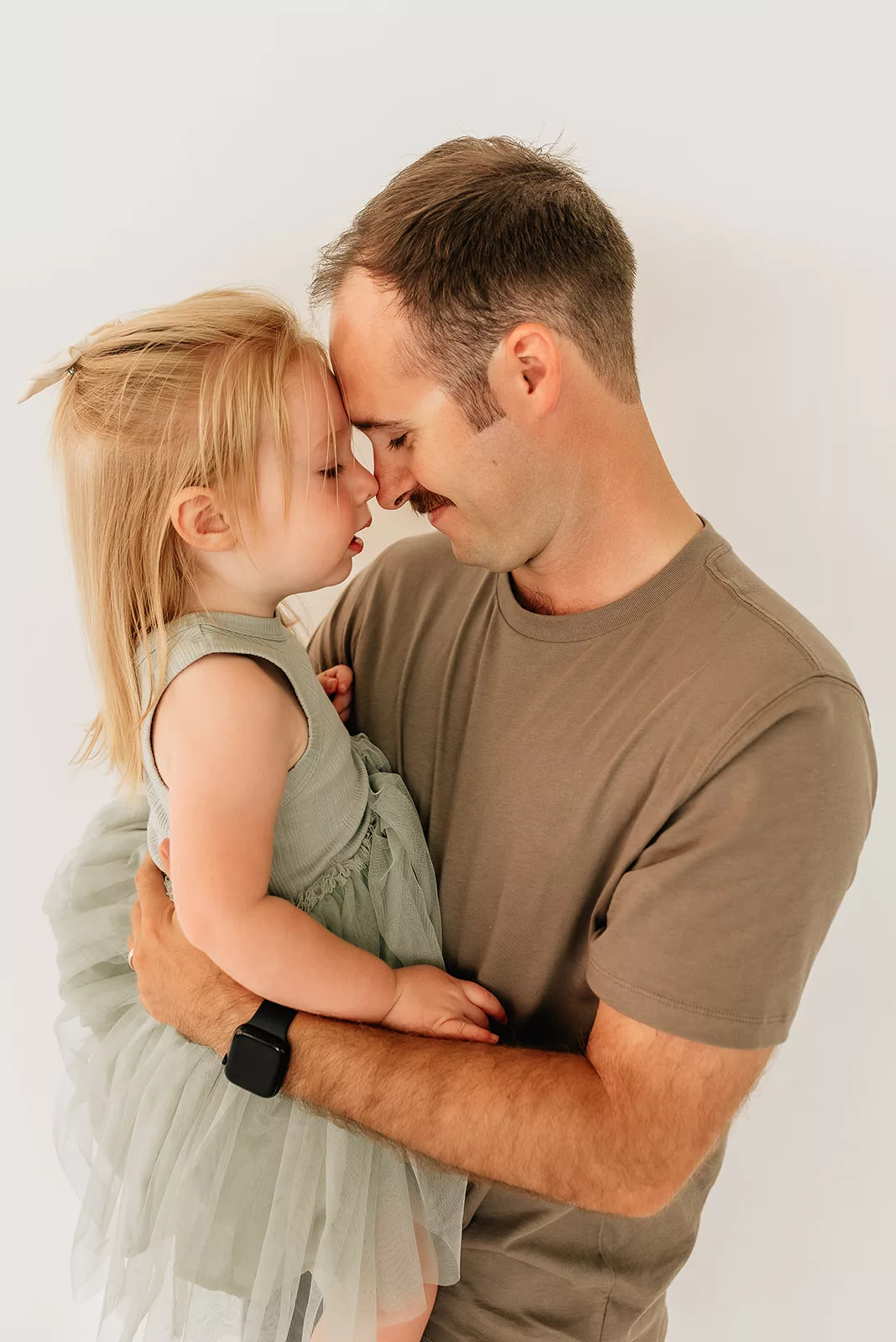 A father nuzzles noses with his toddler daughter sitting in his arms while standing in a studio