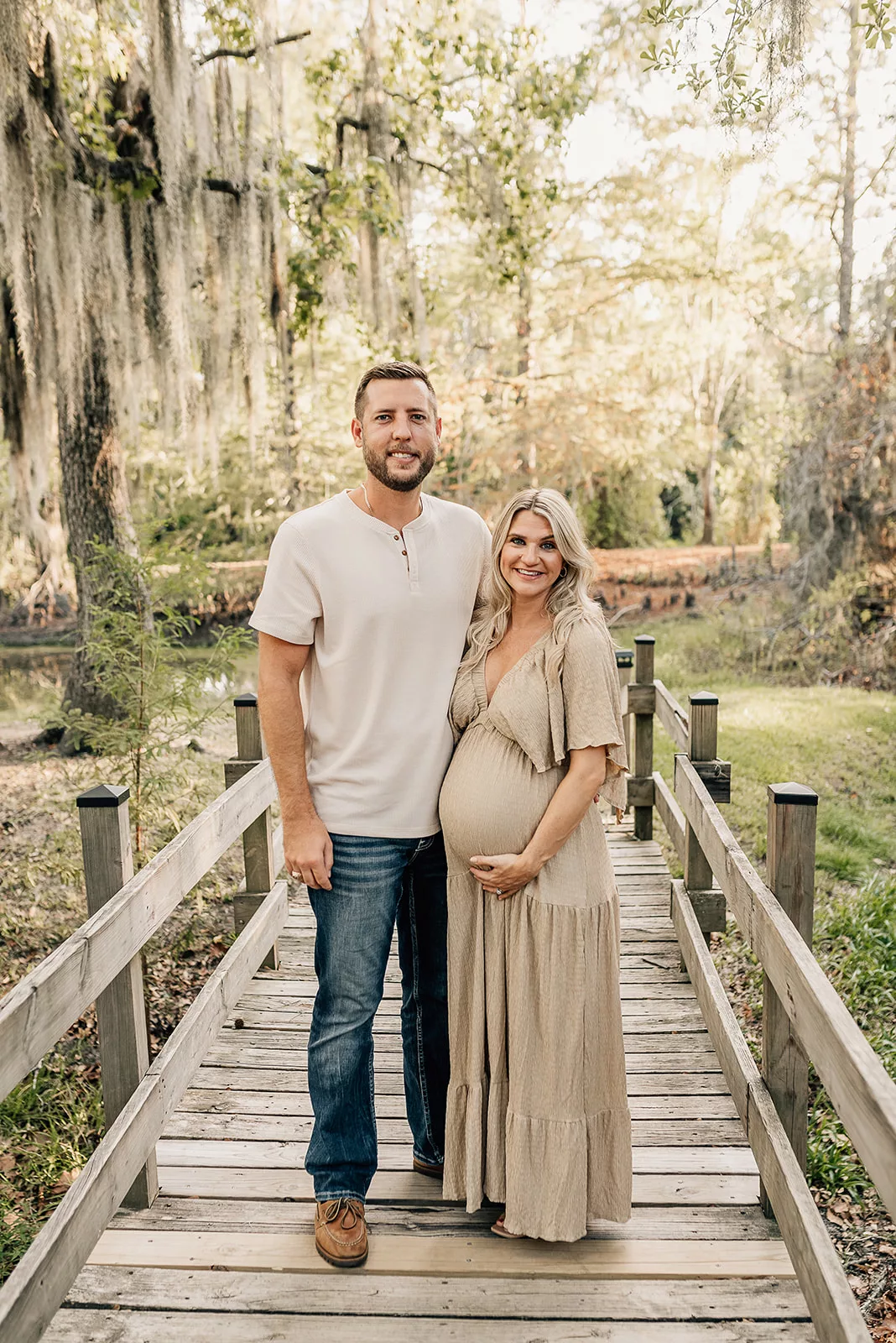 Happy expecting parents stand on a wooden bridge smiling in a park at sunset
