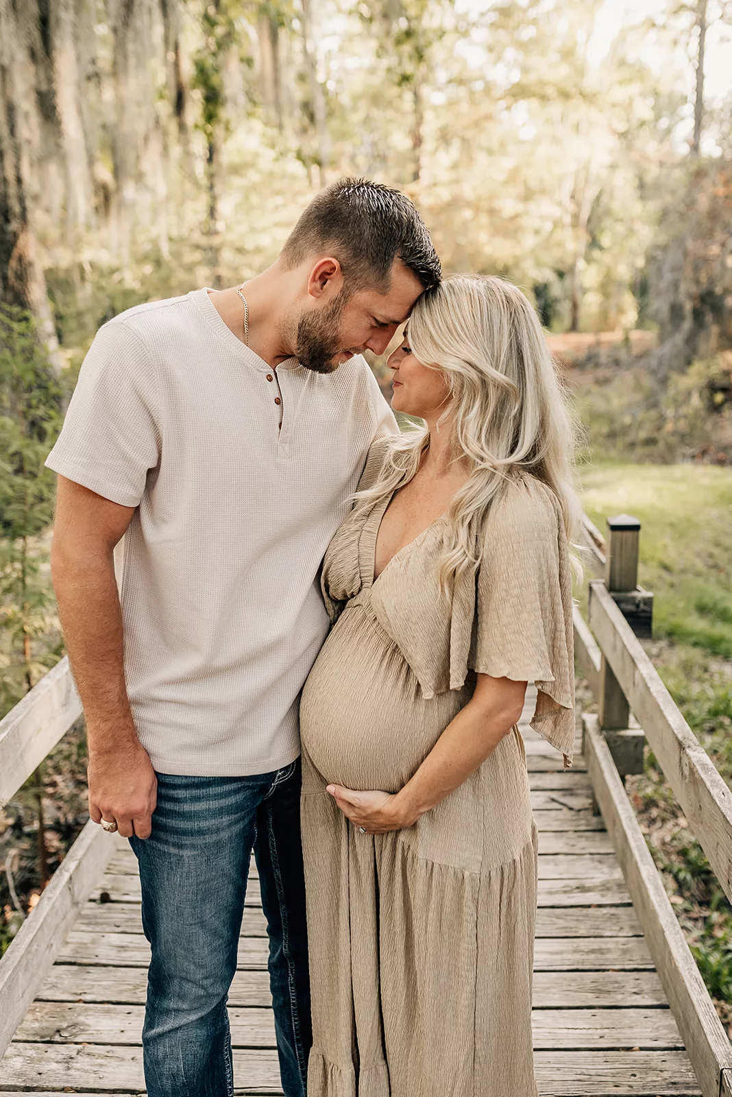 Happy expecting parents place their foreheads together while standing on a wooden bridge with arms around each other
