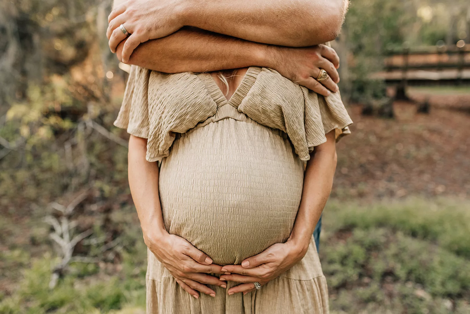 A mom to be in hugged from behind covering her face while holding her bump in a park at sunset