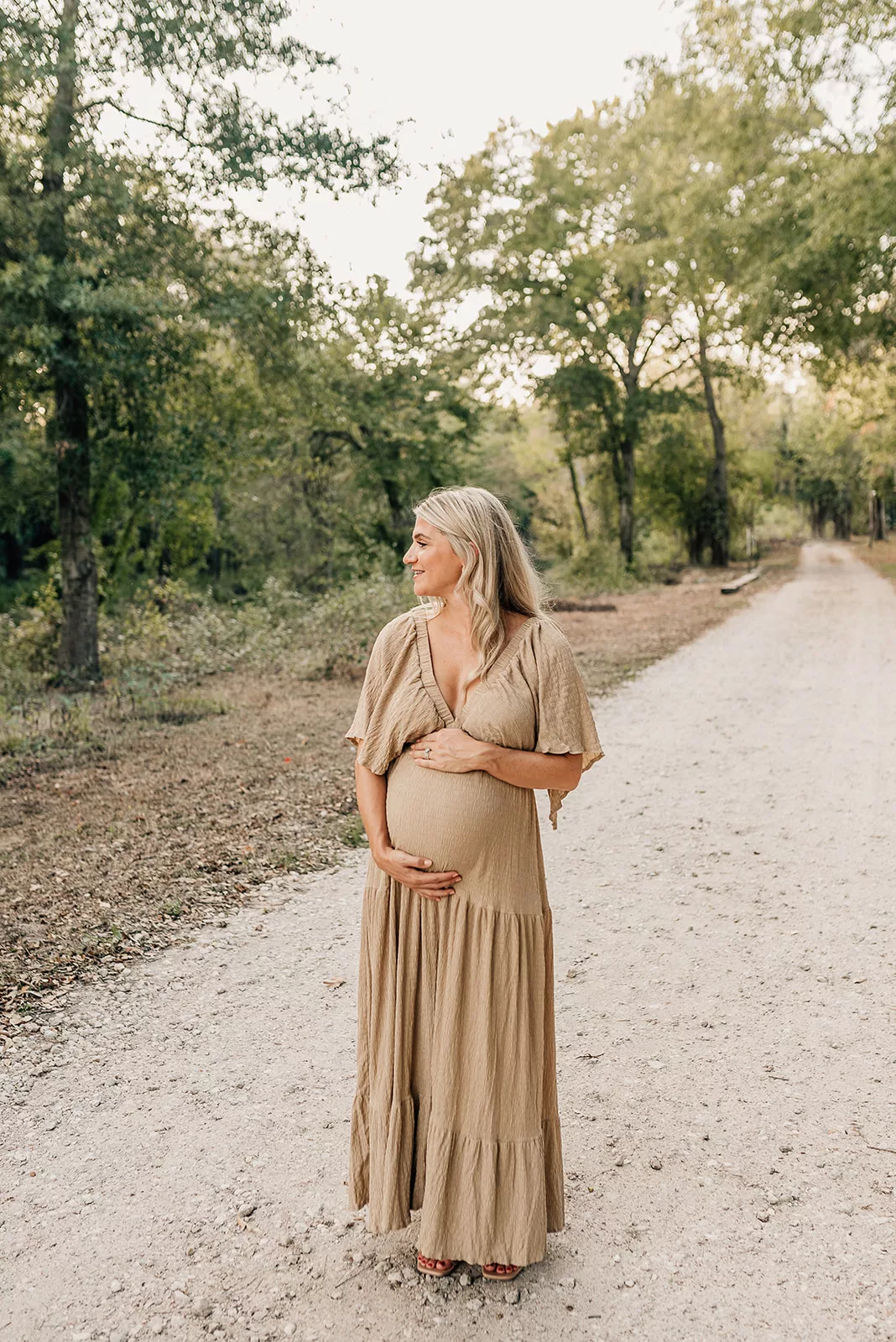 A mother to be happily walks down a park gravel trail while holding her bump in a brown dress
