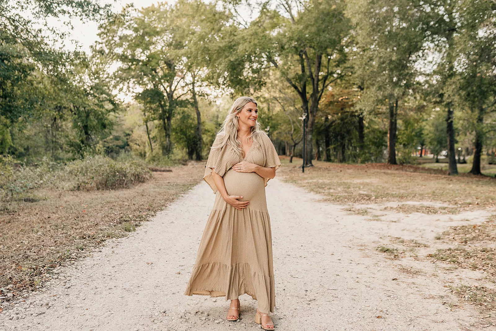 A happy mother to be stands in a park path holding her bump in a beige dress thanks to Houston Fertility Clinics