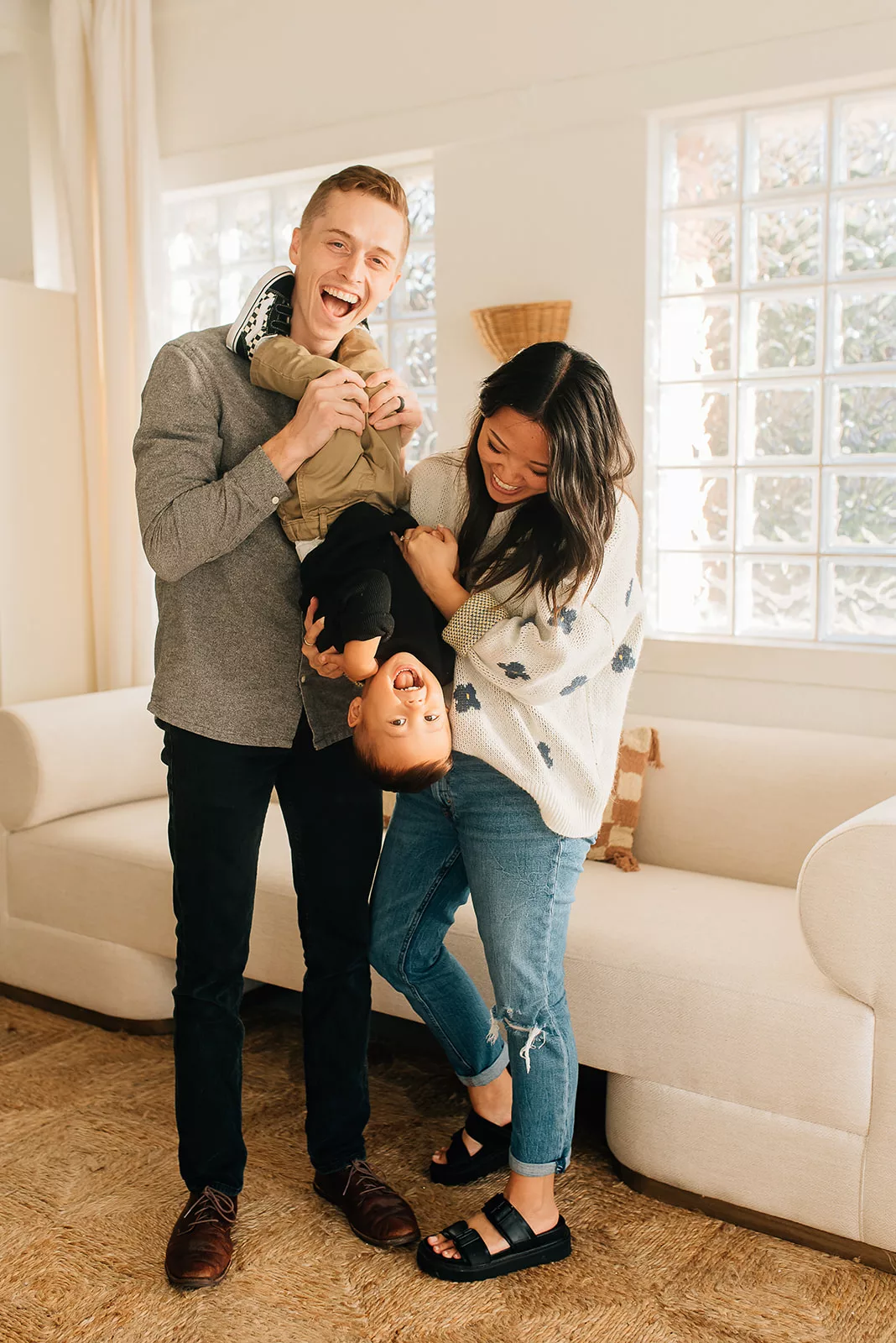 A mother and father tickle and flip over their toddler son in a living room