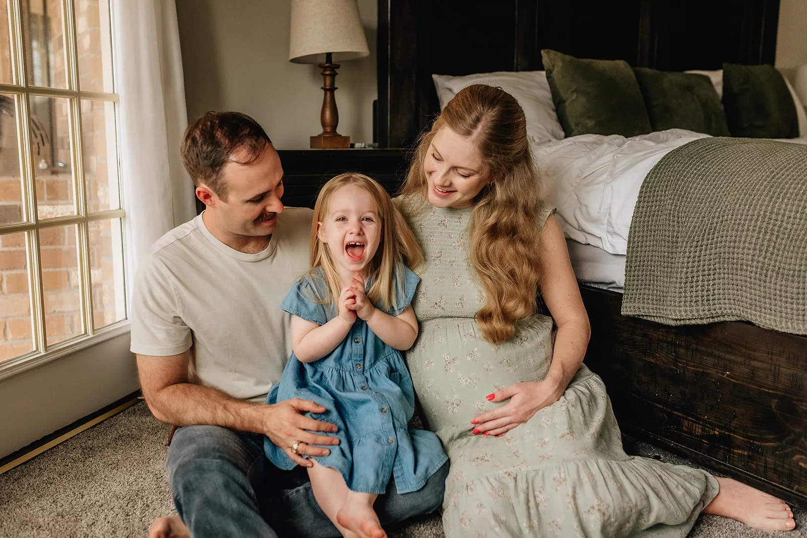 A happy toddler girl claps while sitting in the lap of dad and her pregnant mother