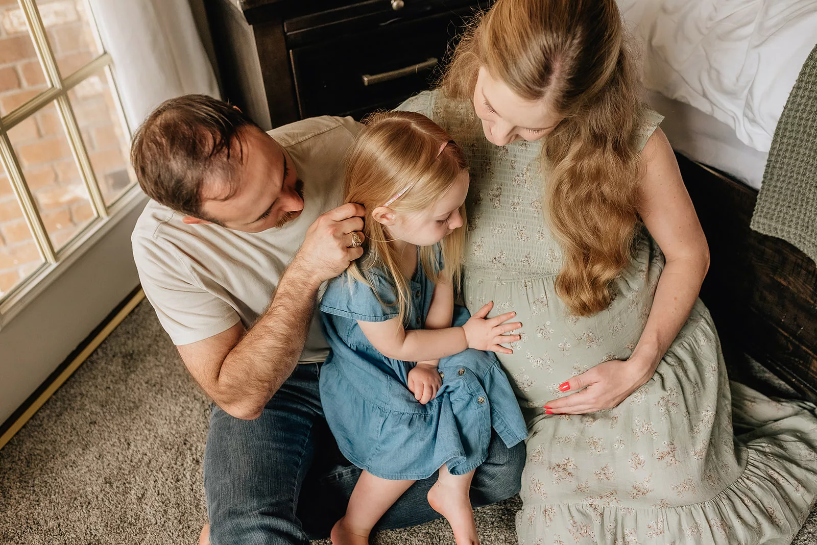 A mom and dad sit on the floor of a bedroom with their toddler daughter touching mom's pregnant bump