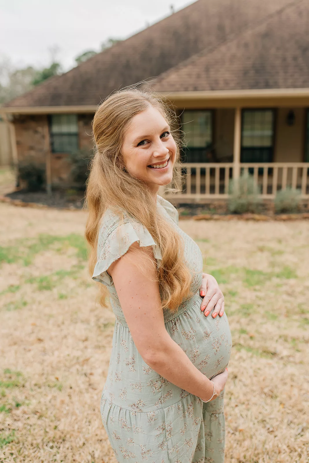 A happy mom to be in a green maternity dress stands smiling over her shoulder in front of a house after meeting a Houston OBGYNs